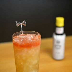 Orchard Punch, an orange-yellow cocktail in a highball glass with pink-orange float of biters at the top with a bow tie cocktail pick