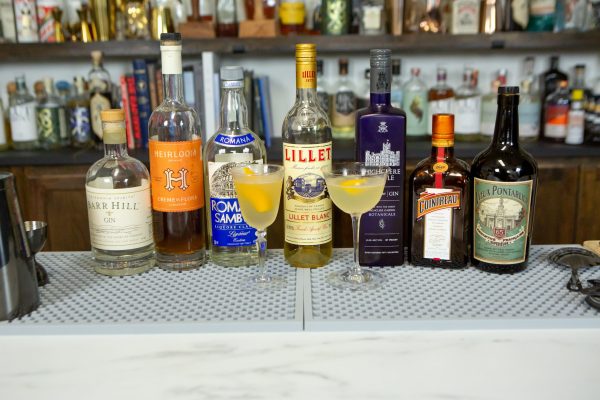 Two Corpse Reviver 2 versions, surrounded by their ingredients and bar tools (shaker, cocktail strainer) on a bar
