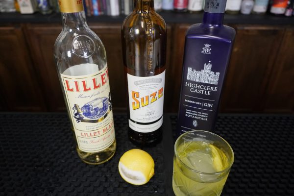 bottles of Lillet, Suze, and gin on a bar next to a White Negroni with a lemon