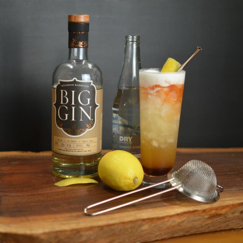 Juniper Pousse cocktail in a tall glass next to a lemon, bottle of gin, and fine strainer