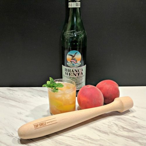 peach cocktail in a rocks glass with 2 peaches, a bottle of alcohol, and a cocktail muddler