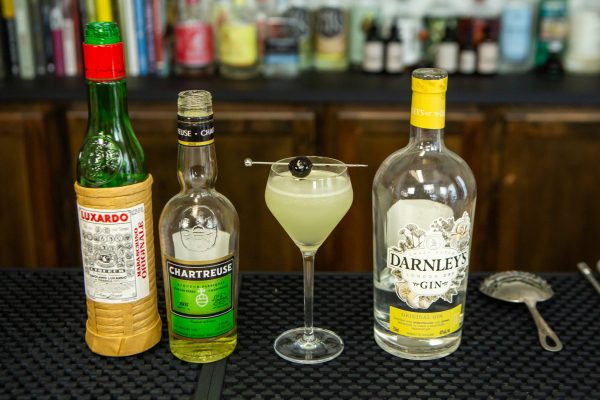 last word cocktail surrounded by luxardo liqueur, chartreuse, and gin bottles plus a julep cocktail strainer