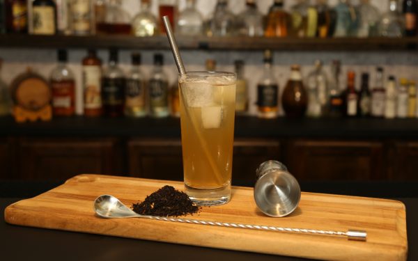 tan cocktail in a collins glass with ice cubes, jigger, bar spoon, and dried flowers
