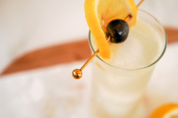 Gold cocktail pick in a sour drink with a fancy cocktail cherry and lemon wheel, in a tall drink glass.