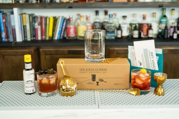 An Old Fashioned cocktail kit with mixing glass, 2 cocktails in rock glass/old fashioned glass, bottle of bitters, jigger, gift box on a bar, making a meaningful gift option