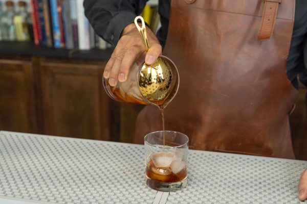 a hand pouring a drink from a mixing glass through julep strainer for weeding out solid ingredients, for a smooth drinking experience
