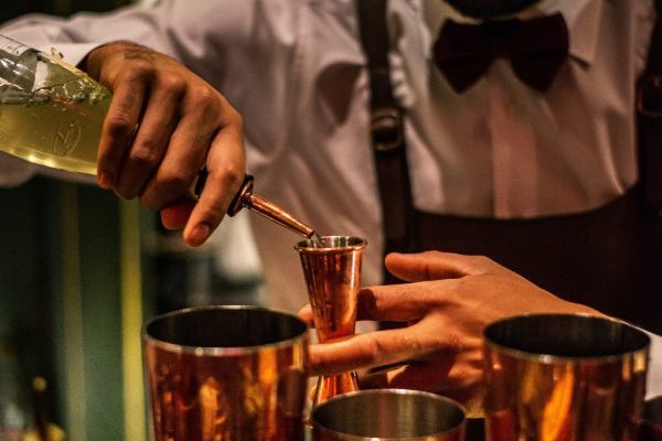 a bartender pouring alcohol into a Japanese jigger in a well-stocked bar with barware around him.