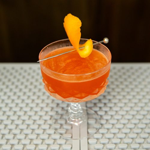Paper Plane drink with an orange twist on a cocktail pick, with lemon juice from a handheld lemon juicer