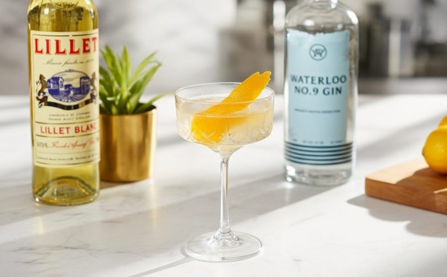 A Vesper cocktail with a lemon peel garnish, next to a bottle of gin and a bottle of Lillet, on a white counter