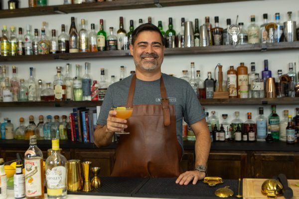 bartender holding a Pegu Club cocktail, with a cocktail shaker and cocktail strainer on the bar next to him