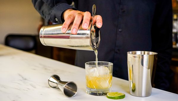 pouring a Margarita from a cocktail shaker into a glass, with stainless steel bar tools on a bar