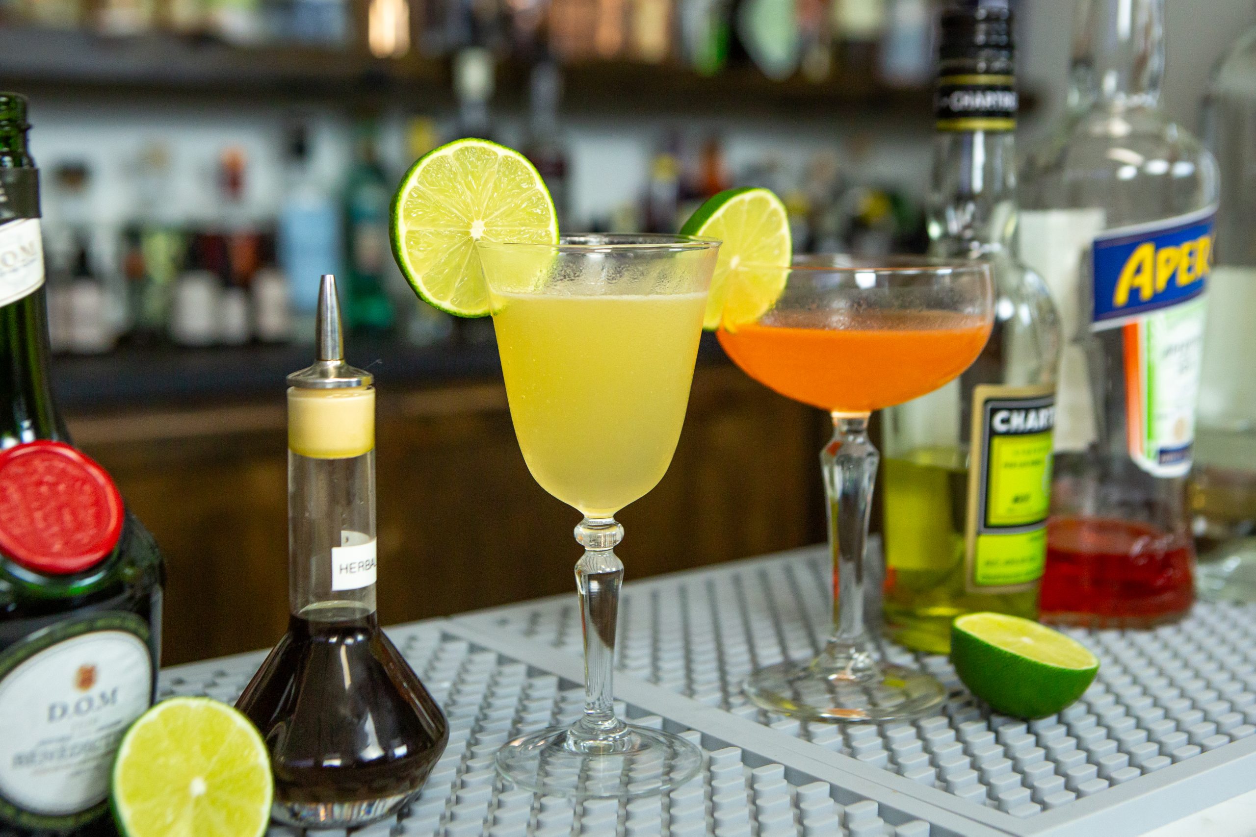 Two cocktails on a bar with ingredients