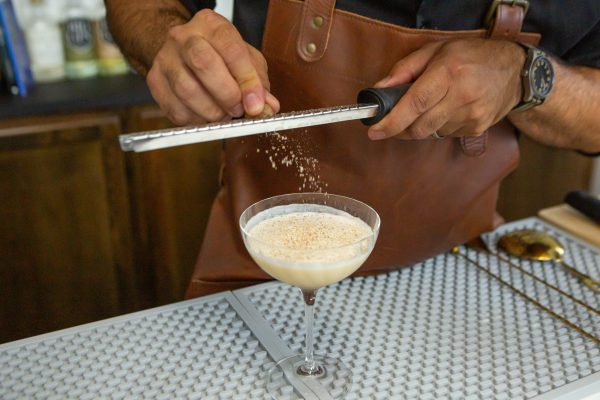 bartender's hand holding a citrus zester, zesting nutmeg over a cocktail