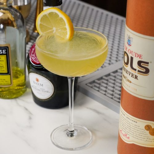 cocktail with lemon wheel, next to its ingredients on a bar