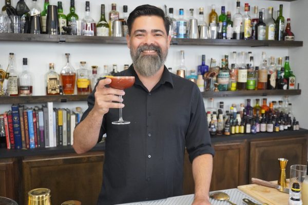 bartender in a bar holding a drink and smiling, surrounded by a cocktail tin, cocktail strainer, and other bar tools and liquor bottles