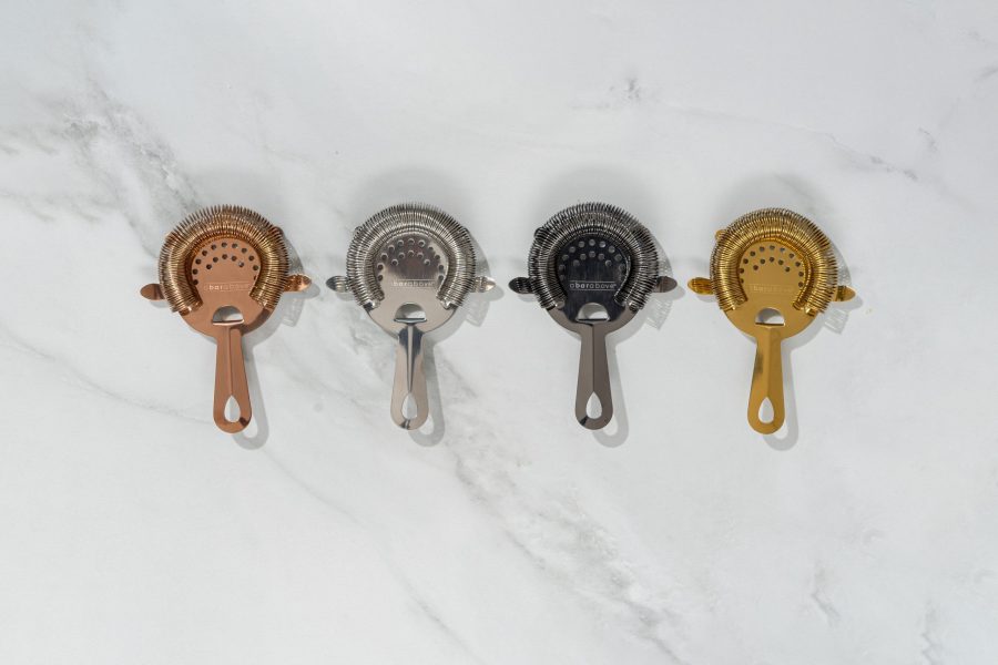 Four 4-pronged Hawthorne cocktail strainers lined up: bronze, silver, black, & gold
