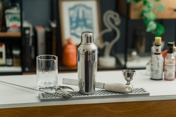 A cobbler shaker, mixing glass, bar spoon, citrus zester, julep strainer, and bell jigger on a bar mat