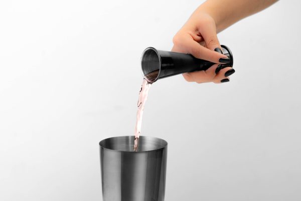 a hand pouring alcohol from a black jigger into a black cocktail shaker