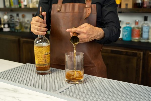 a bartender's hand pouring liquor from a jigger into a cocktail mixing glass