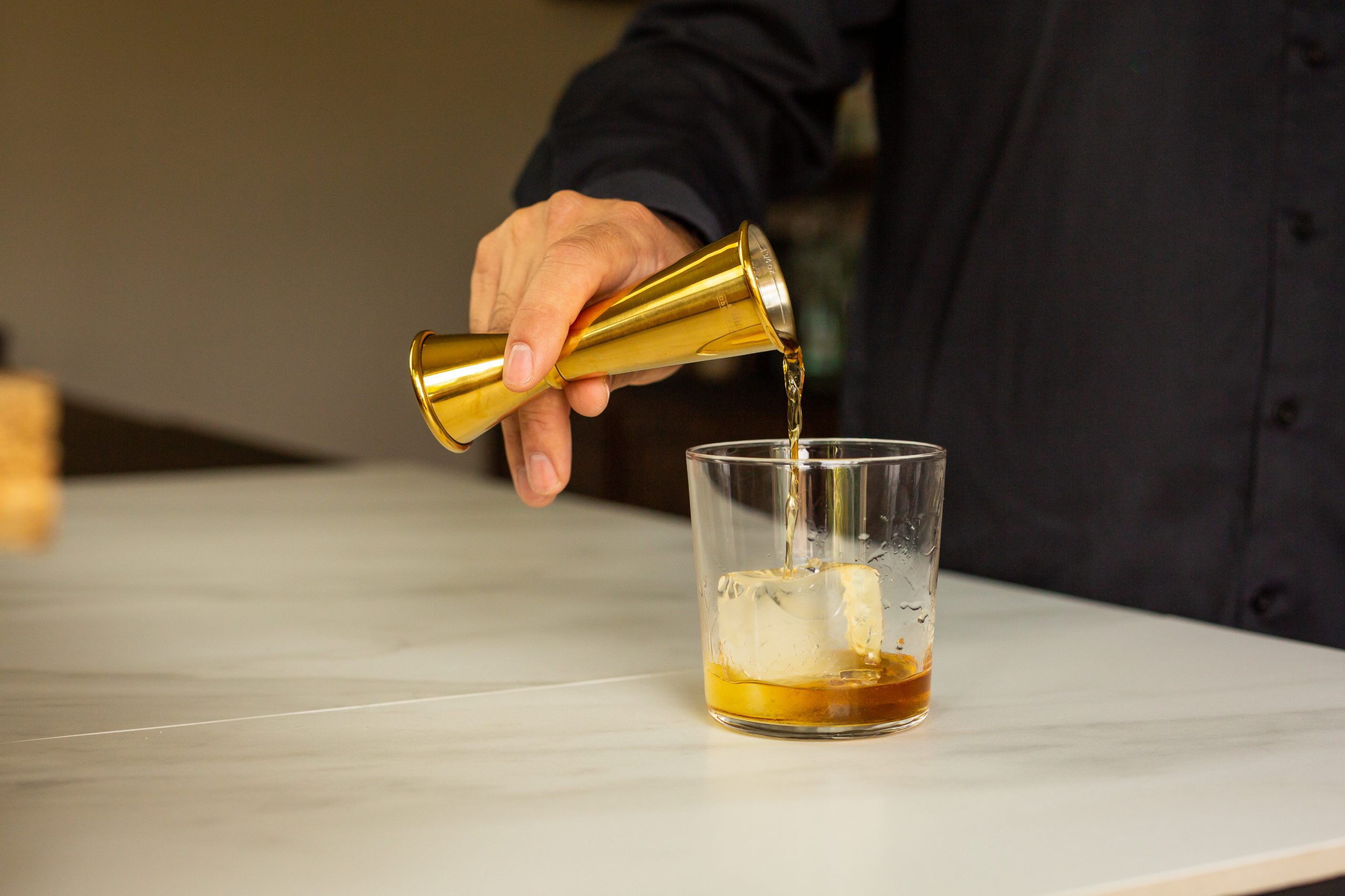a bartender's hand pouring liquor from a gold jigger into a rocks glass with ice