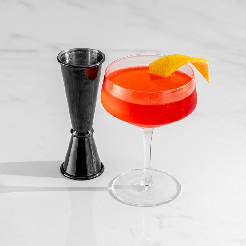 a black Japanese-style cocktail jigger next to a bright red cocktail in a coupe glass