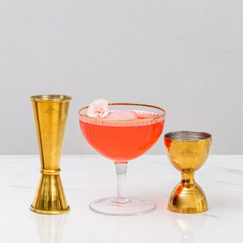 a gold Japanese-style jigger next to a red cocktail in a coupe glass with a gold bell cocktail jigger on the other side
