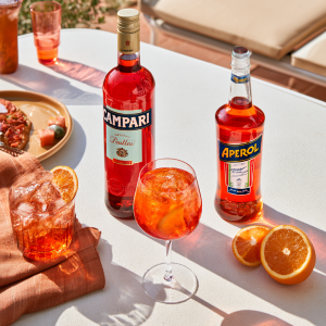 a table set with cocktails and oranges, with a Campari bottle and an Aperol bottle