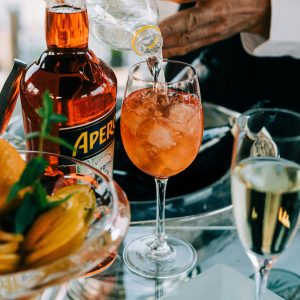 a hand making a cocktail with Aperol in a wine glass with ice, a bottle of amaro next to it