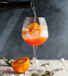 a sparkling wine bottle pouring into a wine glass with an orange and amaro liqueur