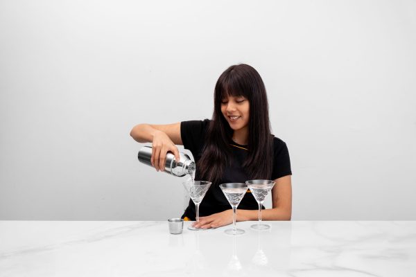 a female bartender pouring a cocktail from a cobbler shaker into a Martini glass