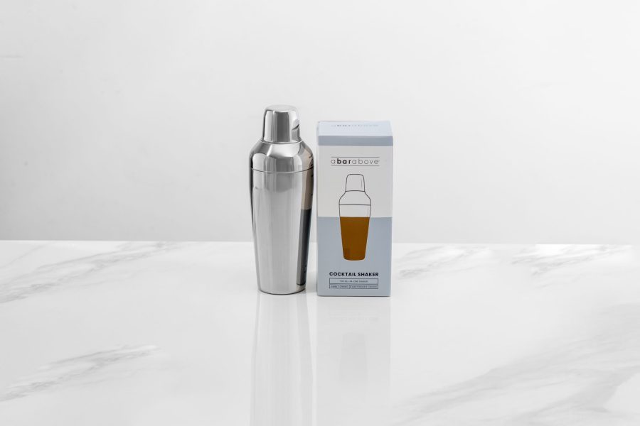 a cobbler style cocktail shaker standing on a marble backdrop next to its gift box