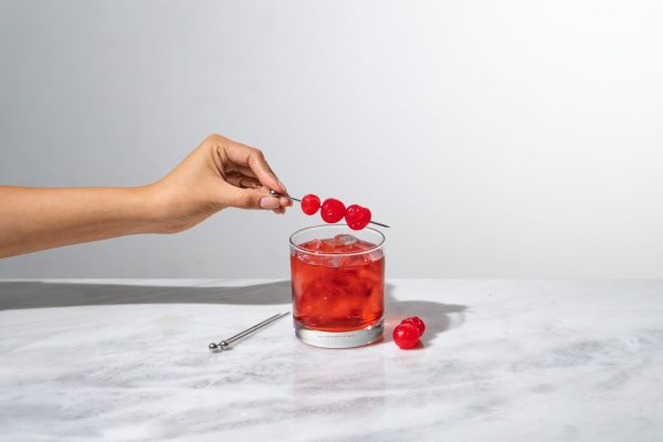 a hand placing a cocktail pick full of cocktail cherries onto a rocks glass (also known as an Old Fashioned glass)