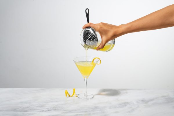 a hand pouring a yellow cocktail into a chilled Martini glass from a mixing glass with a julep cocktail strainer
